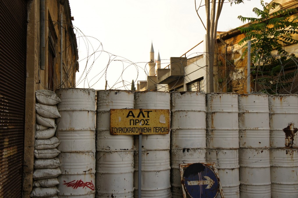 The image shows a barricade made of stacked white barrels and sandbags, topped with barbed wire. In front of the barricade, there is a yellow sign with Greek text and a blue traffic sign with a white arrow pointing to the right. The background includes old buildings and a minaret visible in the distance. The scene appears to be in an urban area with a mix of old and newer structures.