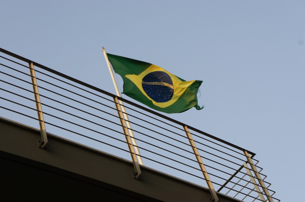The image shows the flag of Brazil waving in the wind. The flag is attached to a pole mounted on a structure with horizontal metal railings. The sky in the background is clear and blue.