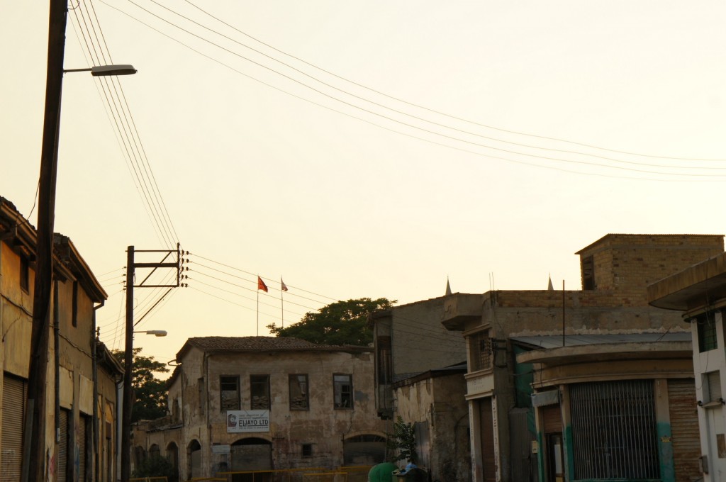 The image shows an urban street scene at sunset or early evening. The buildings appear to be old and somewhat dilapidated, with visible wear and tear. There are power lines and a streetlight on the left side of the image. In the background, there are a few flags on poles, and some trees are visible behind the buildings. The sky is a pale, warm color, indicating the time of day. One of the buildings has a sign that reads "ELJAYO LTD."