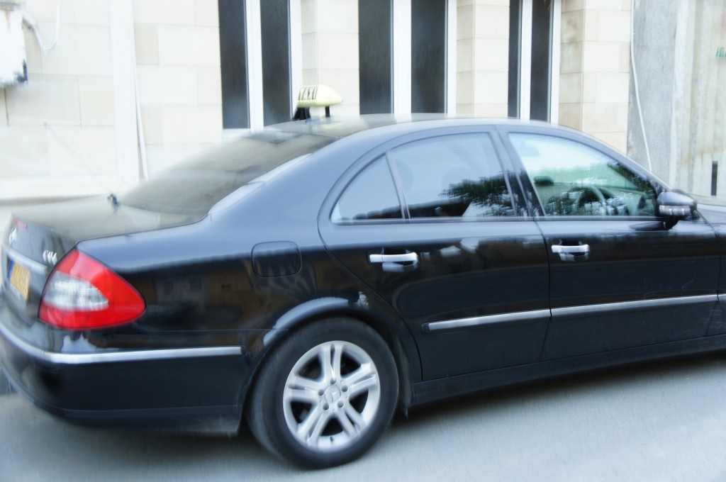 A black sedan taxi is parked on the side of the street in front of a building with beige stone walls and vertical black and white stripes on the windows. The taxi has a yellow sign on its roof. The image is slightly blurred.