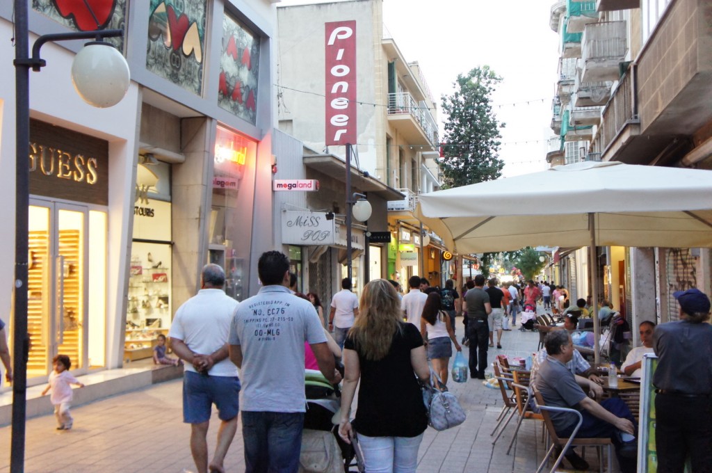 A bustling pedestrian street lined with shops and cafes. People are walking along the street, some carrying shopping bags, while others are seated at outdoor tables under large umbrellas. The shops have various signs, including one for "GUESS" and another for "Pioneer." The street is lively with activity, and the buildings are multi-story with balconies. There are also some decorative lights strung across the street.
