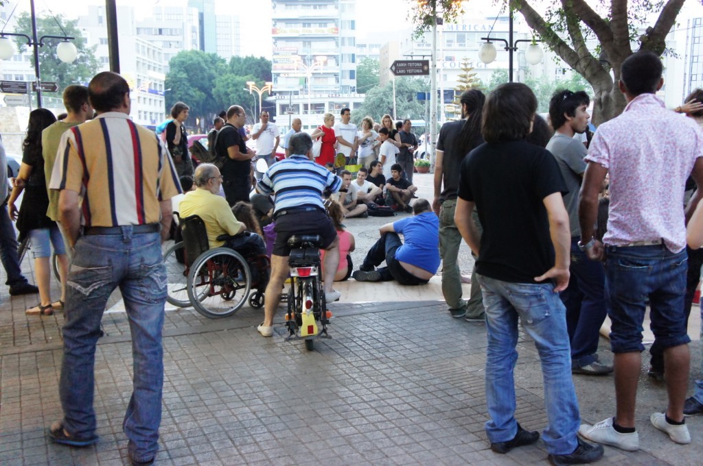 A group of people is gathered in an outdoor urban area. Some individuals are standing, while others are sitting or kneeling on the ground. One person is in a wheelchair, and another is on a bicycle. The background features buildings, trees, and street lamps. The atmosphere appears to be casual, possibly a public gathering or event.