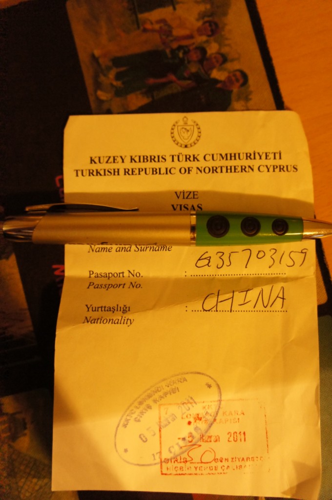 The image shows a visa document from the Turkish Republic of Northern Cyprus. The document includes fields for "Name and Surname," "Passport No.," and "Nationality." The passport number is handwritten as "G35703157," and the nationality is listed as "CHINA." There is a pen placed on top of the document. The document also has a stamp dated "30 Haziran 2011" (June 30, 2011). In the background, there is a partially visible photograph of a group of people.