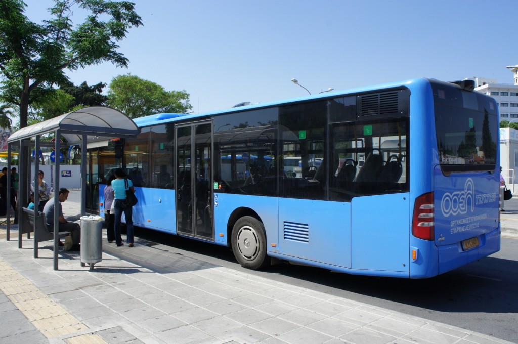 A blue city bus is parked at a bus stop. Several people are waiting at the bus stop, with some standing and others sitting on benches under a covered shelter. The bus has large windows and a door open for passengers to board. Trees and buildings are visible in the background under a clear blue sky.