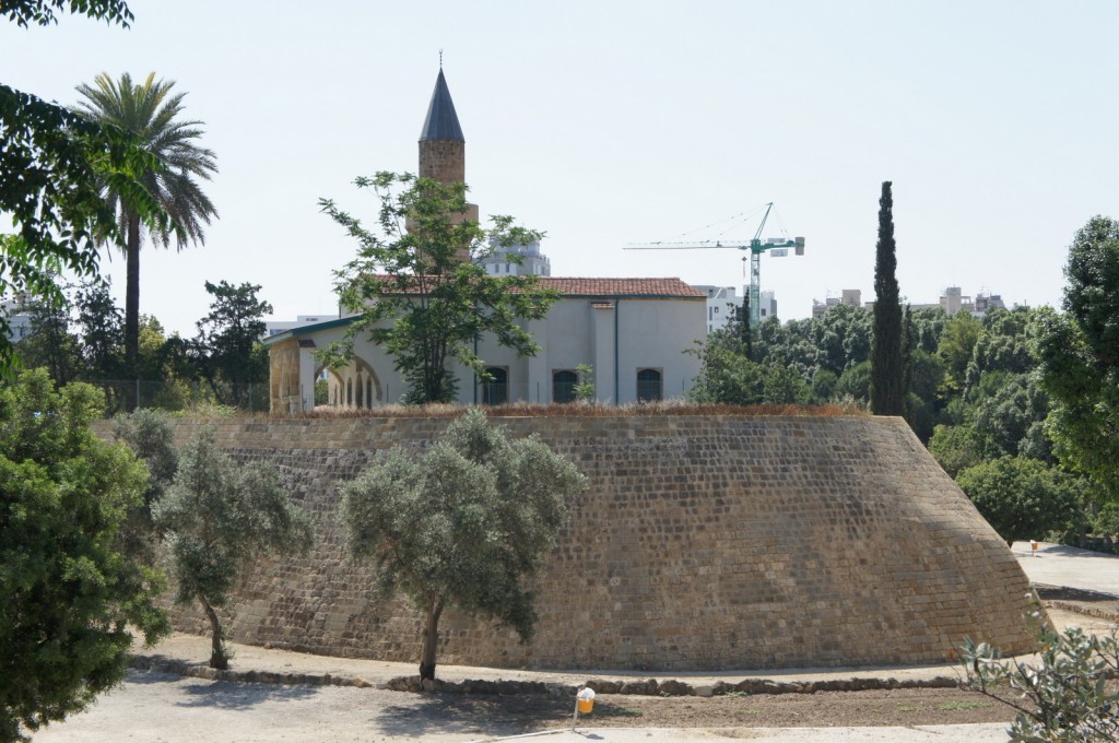 The image shows a historic building with a tall, pointed tower, surrounded by lush greenery. The building is situated on a raised, stone-walled platform. There are various trees, including a palm tree and other types of foliage, around the structure. In the background, a construction crane and modern buildings are visible, indicating an urban setting. The sky is clear and bright, suggesting a sunny day.
