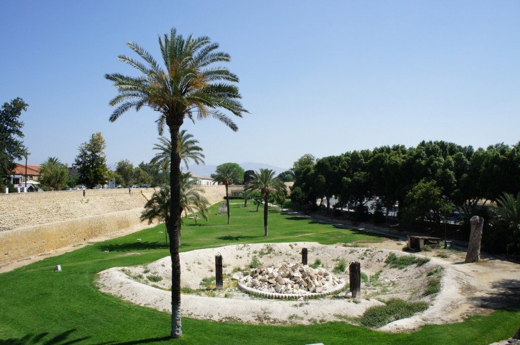 The image shows a scenic outdoor area with a mix of natural and historical elements. In the foreground, there is a circular excavation site filled with rocks and surrounded by a low barrier. Several tall palm trees are scattered throughout the grassy area, providing a tropical feel. To the left, there is a stone wall running parallel to a pathway, with some buildings and more trees visible in the background. On the right side, a dense line of trees creates a natural border. The sky is clear and blue, indicating a sunny day.