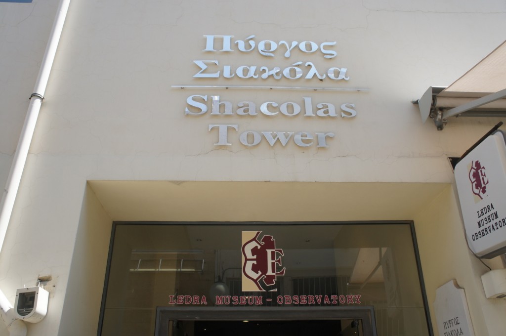 The image shows the entrance to the Shacolas Tower, with the name displayed in both Greek and English. Below the name, there is a sign for the Ledra Museum and Observatory. The building has a light-colored exterior, and there is a sign on the right side of the entrance with the same information.