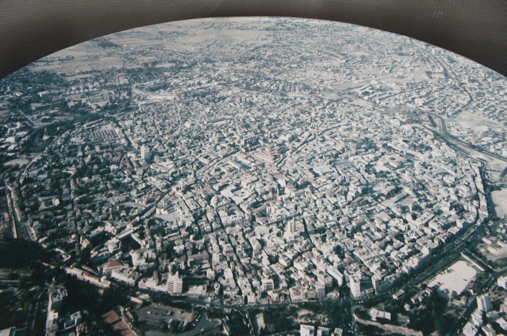 Aerial view of a densely populated urban area with numerous buildings, streets, and some green spaces. The cityscape extends into the distance, showing a mix of residential, commercial, and possibly industrial areas. The image is taken from a high vantage point, providing a broad perspective of the city's layout and infrastructure.