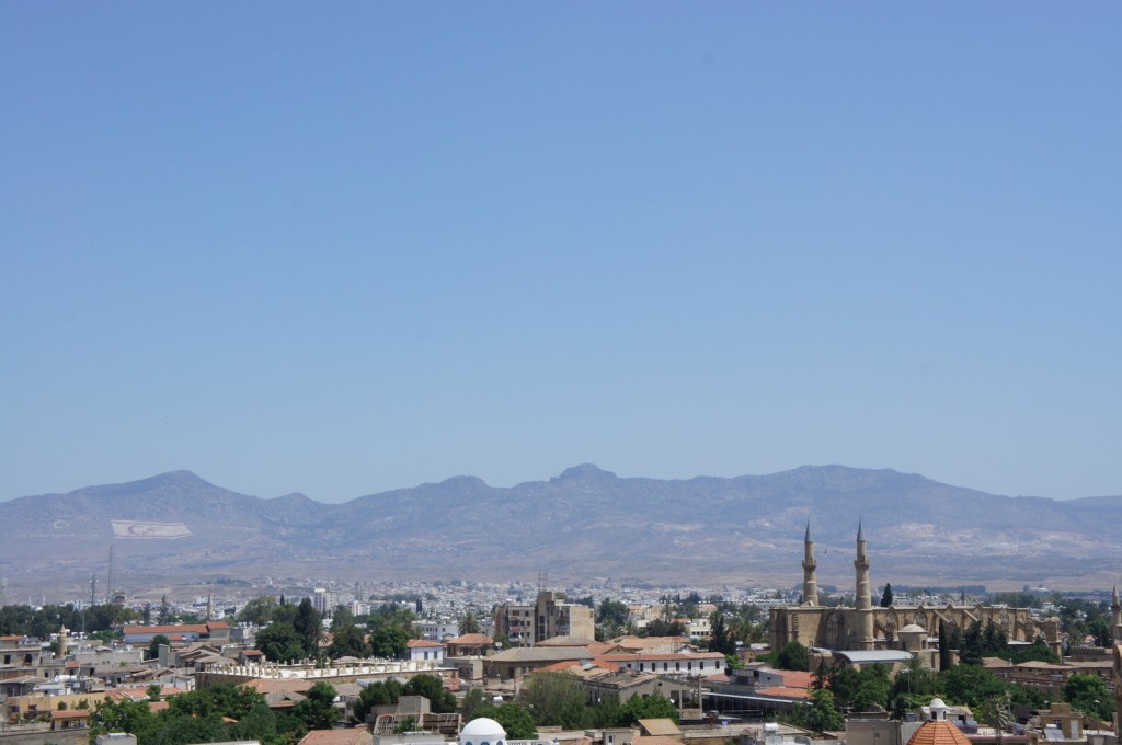 The image shows a cityscape with a mix of modern and traditional buildings, including a prominent mosque with two tall minarets. In the background, there are mountains with a clear blue sky above. The city appears to be situated in a valley or flat area with the mountains rising behind it. The overall scene is bright and sunny.