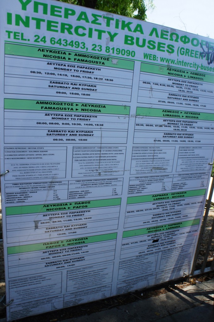 The image shows a bus schedule board for Intercity Buses in Cyprus. The text is in both Greek and English. The board lists various routes, including Nicosia to Famagusta, Famagusta to Nicosia, Nicosia to Limassol, Limassol to Nicosia, Nicosia to Paphos, Paphos to Nicosia, Larnaca to Nicosia, and Nicosia to Larnaca. Each route has specific departure times for Monday to Friday and for Saturday and Sunday. The board also includes contact telephone numbers and a website URL for more information.