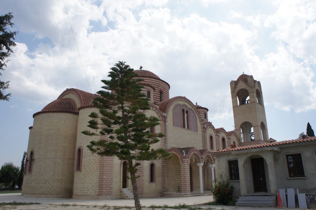 The image shows a large, beige-colored building with red-tiled roofs, featuring architectural elements typical of a church, such as arched windows and doorways. The structure includes a prominent bell tower on the right side and multiple domes. In the foreground, there is a tall, conical tree, and the sky above is partly cloudy.