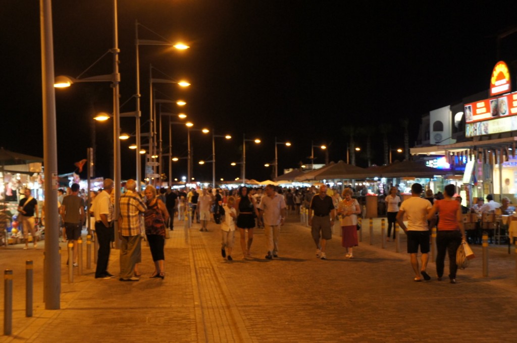 A bustling street scene at night with numerous people walking and socializing. The street is well-lit with a series of street lamps, and there are various shops and restaurants on either side. Some people are sitting at outdoor tables, while others are browsing or walking along the street. The atmosphere appears lively and vibrant.