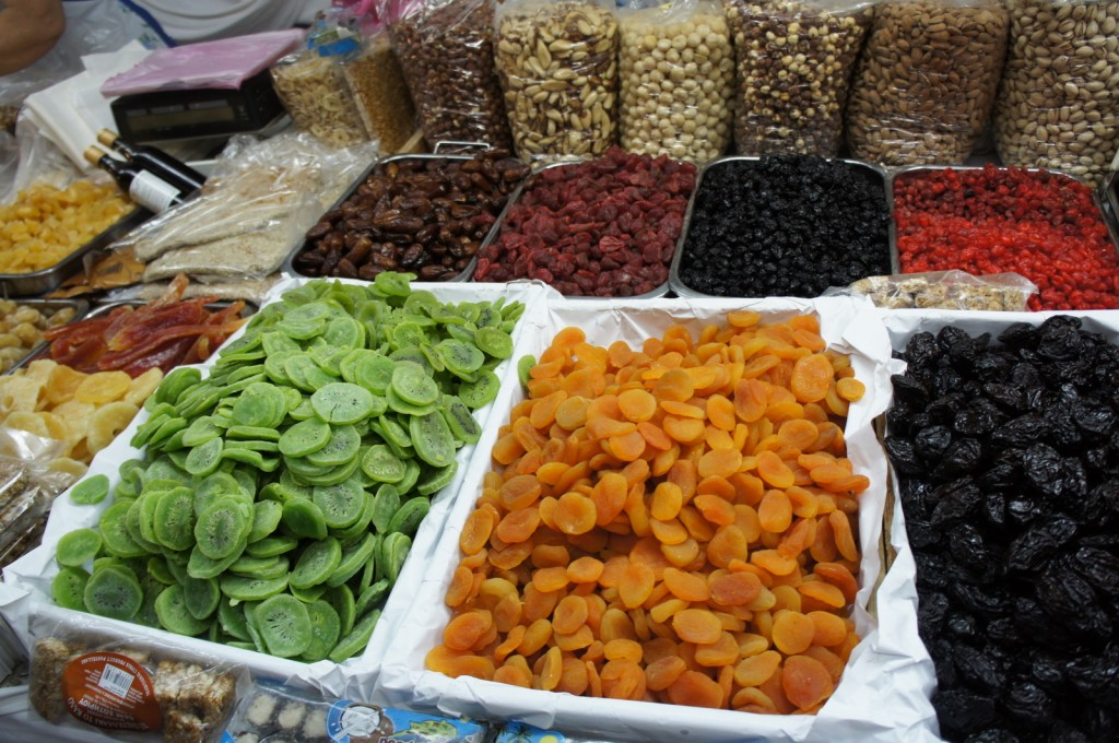 The image shows a market stall displaying a variety of dried fruits and nuts. In the foreground, there are trays filled with dried green kiwi slices, orange apricots, and dark prunes. Behind these trays, there are containers with different types of dried fruits such as dates, red berries, and blackberries. In the background, there are bags filled with various nuts including almonds, pistachios, and hazelnuts. The stall also has some packaged goods and bottles of liquid on the left side.
