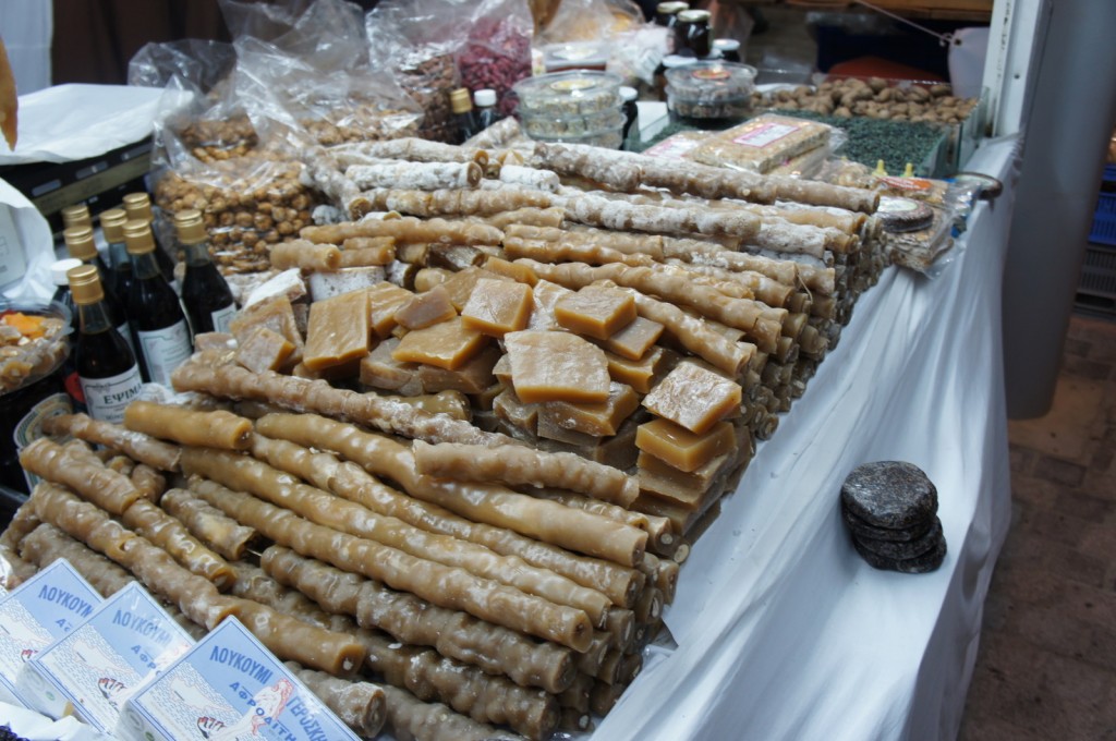 The image shows a market stall displaying a variety of traditional sweets and food items. There are numerous long, cylindrical sweets that appear to be coated in powdered sugar, possibly a type of nougat or nut-filled candy. In the center, there are square pieces of a caramel-colored confection. Surrounding these items are bottles of what seems to be wine or another type of beverage, and various packaged nuts and dried fruits. The stall is covered with a white tablecloth, and the items are neatly arranged for display.