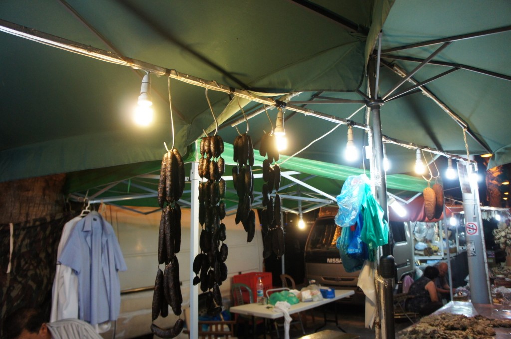 A market stall is shown under a green canopy, illuminated by several hanging light bulbs. Various items, including sausages and other food products, are hanging from the canopy's frame. There are also some plastic bags and clothing items visible. In the background, there is a table with various items on it, and a van parked behind the stall. The scene appears to be set in the evening or night.