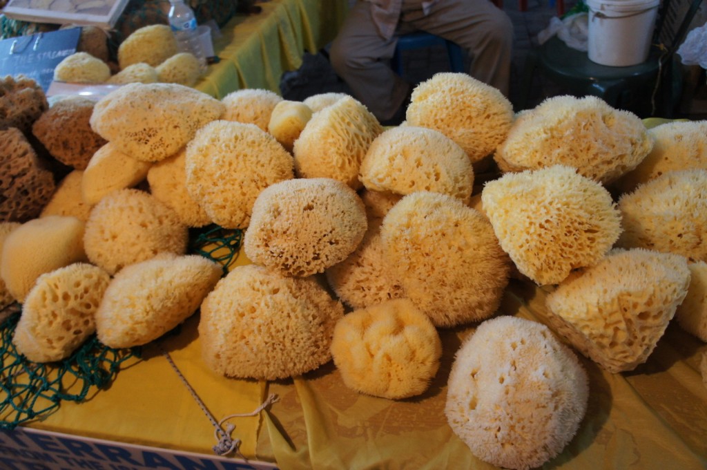 The image shows a display of natural sea sponges arranged on a table. The sponges are various shades of yellow and beige, with a porous texture. They are piled together, and the table is covered with a yellow cloth. In the background, there are more sponges and some other items, including a water bottle and a white bucket.