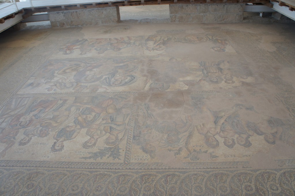 Alt text: The image shows an ancient mosaic floor with intricate designs and depictions of human figures. The mosaic appears to be partially damaged or worn, with some sections more faded than others. The artwork is enclosed by a border with geometric patterns. The scene is housed in an archaeological site, with a protective structure overhead.