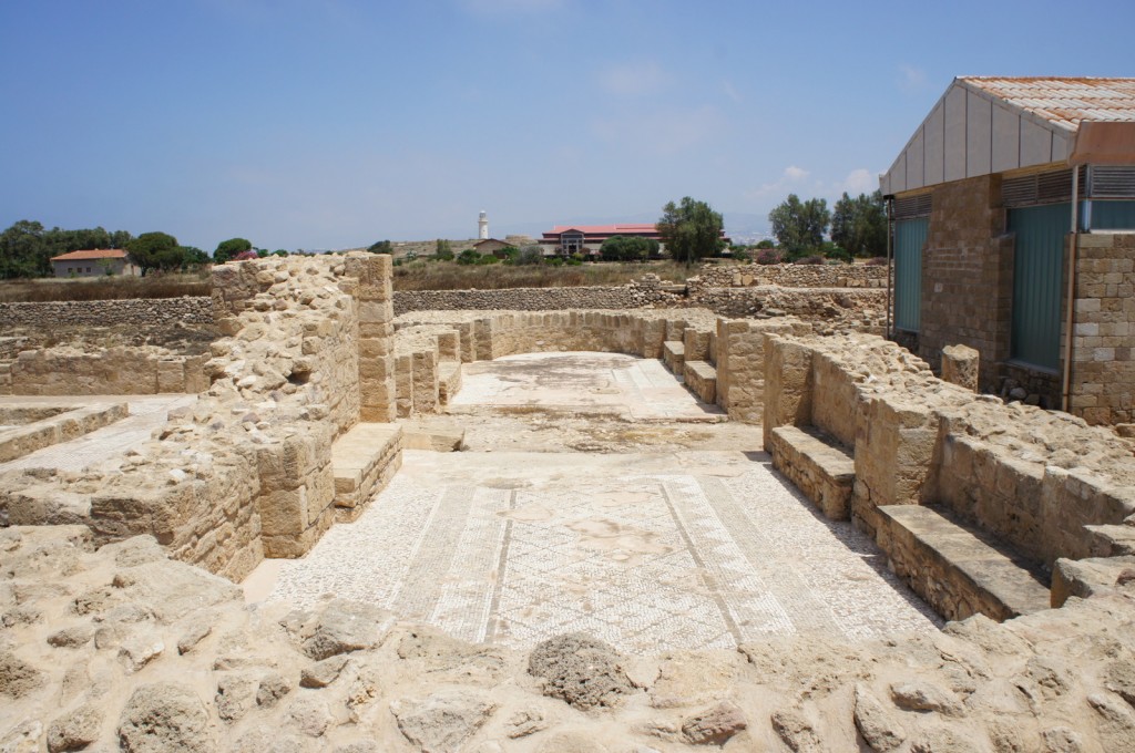 Alt text: The image shows ancient ruins with partially standing stone walls and mosaic floors. The site appears to be an archaeological excavation area. In the background, there are modern buildings and a lighthouse, with trees and a clear blue sky.