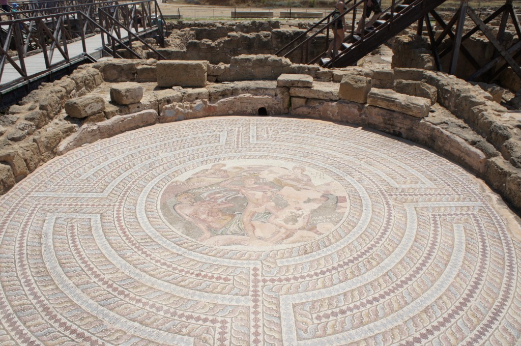 The image shows an ancient circular mosaic floor with intricate geometric patterns and a central depiction of human figures. The mosaic is surrounded by stone ruins, and there are wooden walkways and railings around the site. In the background, a few people are seen walking on the wooden walkways. The scene appears to be an archaeological site, possibly an ancient Roman or Greek ruin.