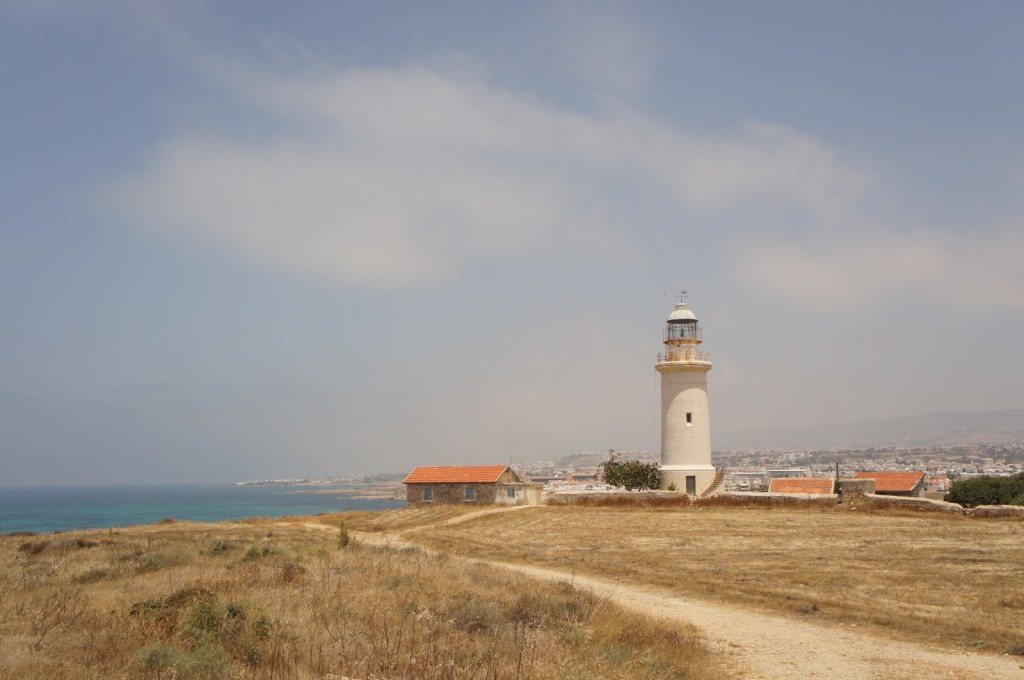 The image shows a coastal landscape with a lighthouse and a small building with a red-tiled roof. The lighthouse is white and cylindrical, standing tall on a grassy, dry field. In the background, there is a view of the sea on the left and a distant town or city with more buildings and houses. The sky is partly cloudy with a light blue hue.