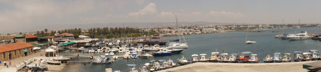 Panoramic view of a bustling marina with numerous boats and yachts docked along the piers. The marina is surrounded by various buildings, including some with red-tiled roofs. In the background, there is a cityscape with a mix of low-rise and mid-rise buildings, and a range of hills or mountains can be seen in the distance under a partly cloudy sky. The water is calm, and the overall scene suggests a lively coastal area.