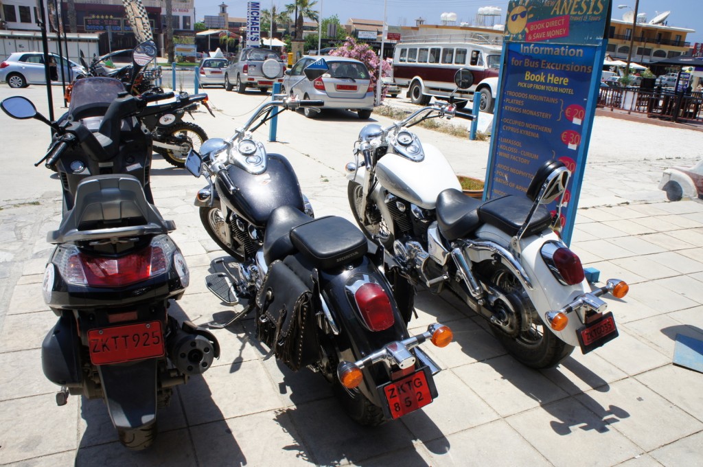 The image shows a row of parked motorcycles and scooters on a paved area. There are four vehicles in the foreground, with two black motorcycles, one white motorcycle, and one black scooter. The motorcycles have red license plates. In the background, there are more vehicles, including cars and a bus, as well as a signboard providing information about bus excursions. The area appears to be a parking lot or a public space near a tourist information center. There are buildings, trees, and other signs visible in the background.