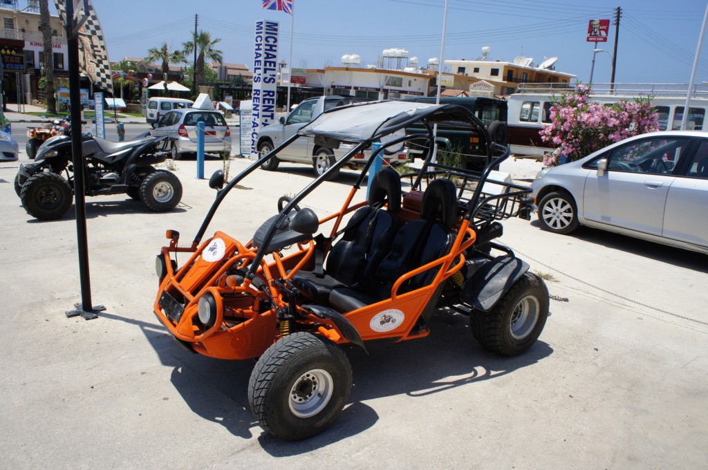 The image shows an orange off-road dune buggy parked in a lot. The buggy has two seats and a roll cage. In the background, there is a black ATV (all-terrain vehicle) and several other vehicles, including a silver car. There are also signs and buildings in the background, one of which has a banner that reads "MICHAEL'S RENT-A-CAR." The sky is clear and blue, indicating a sunny day.