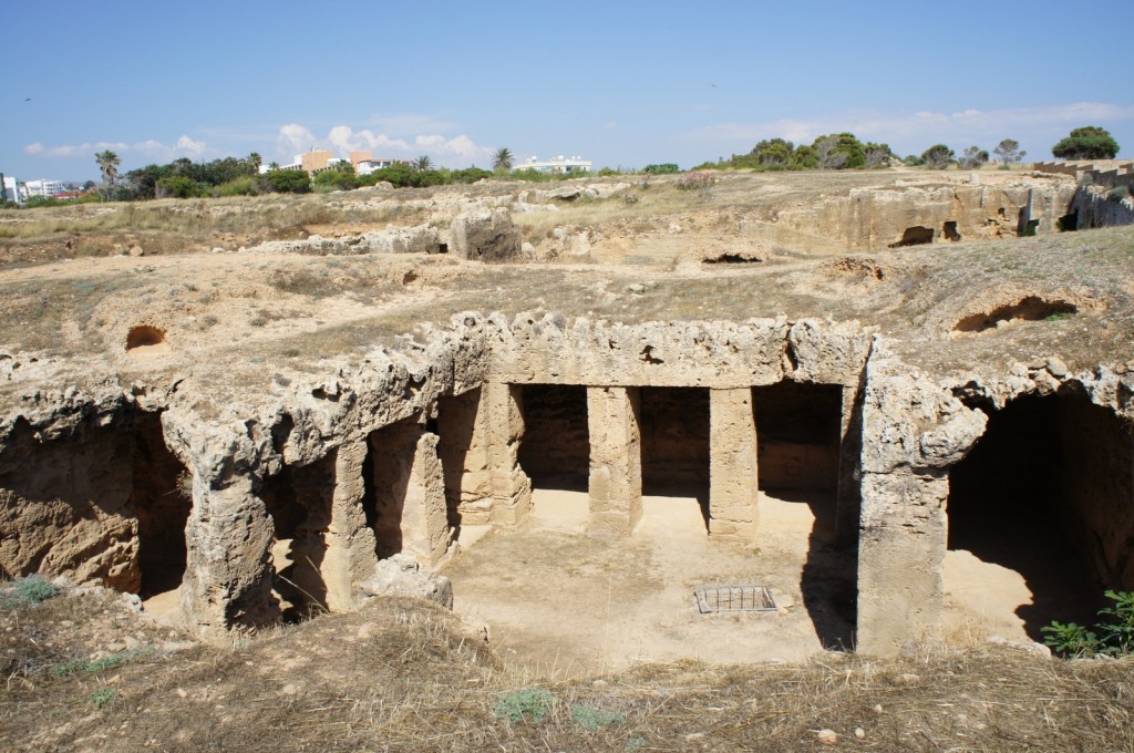 The image shows an ancient archaeological site with rock-cut structures. The site appears to be a series of interconnected chambers or rooms carved into the ground, with some areas supported by stone pillars. The terrain is dry and rocky, with sparse vegetation. In the background, there are some modern buildings and trees, indicating that the site is located near an urban area. The sky is clear and blue, suggesting a sunny day.