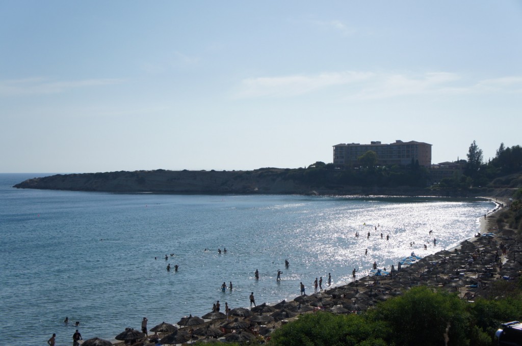 A scenic view of a beach with people swimming and enjoying the water. The shoreline is lined with numerous beach umbrellas and sunbathers. In the background, there is a large building, possibly a hotel or resort, situated on a hill overlooking the beach. The sky is clear, and the sun is reflecting off the water, creating a shimmering effect.