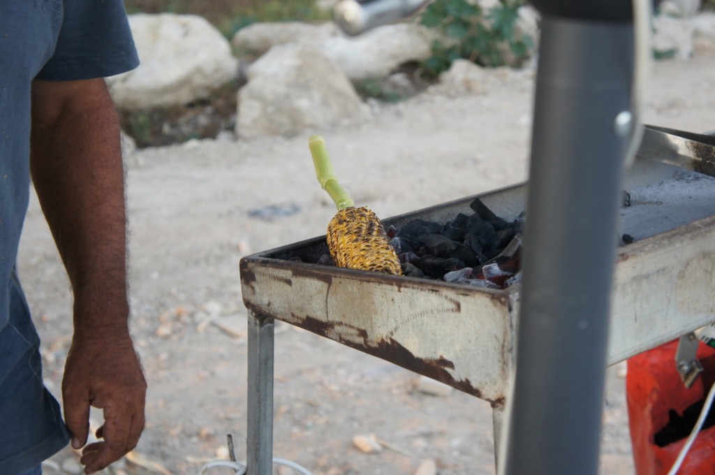 A person is standing next to a metal grill with a piece of corn on the cob being roasted over charcoal. The background is outdoors with some rocks and greenery visible.