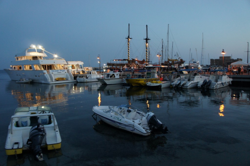 A marina at dusk with various boats docked. The scene includes a large, well-lit yacht named "Ocean Flyer" on the left, several smaller boats in the middle, and a few more boats on the right. The water reflects the lights from the boats and the dock, creating a serene atmosphere. The sky is clear, transitioning from light to dark as evening sets in.