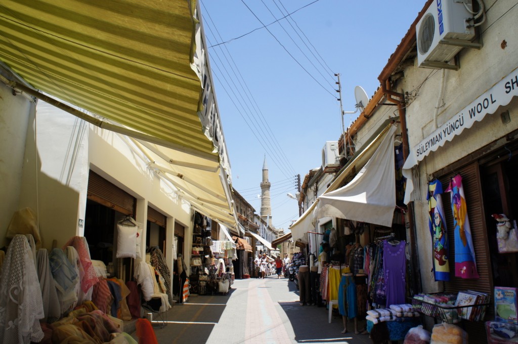A bustling outdoor market street with various shops on both sides. The shops have awnings providing shade, and they display a variety of goods including fabrics, clothing, and other textiles. There are signs in a foreign language, and one shop sign reads "S&uuml;leyman Y&uuml;nc&uuml; Wool Shop." In the background, a tall minaret is visible, indicating the presence of a mosque. The sky is clear and blue, suggesting a sunny day. People can be seen walking and shopping along the street.
