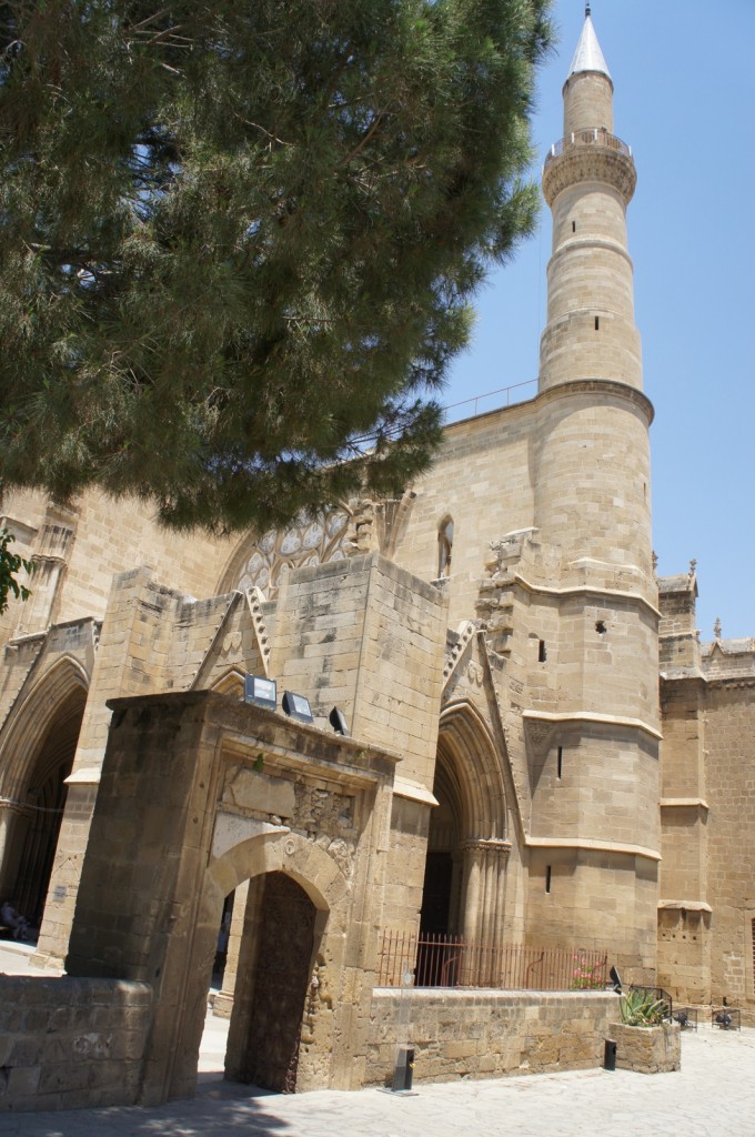 Alt text: A historic stone building with a tall minaret and arched doorways. The structure features Gothic architectural elements and is partially obscured by a large tree in the foreground. The sky is clear and blue.