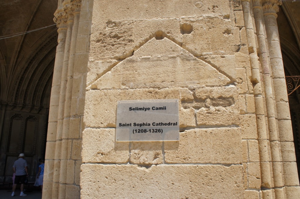 The image shows a close-up of a stone wall with a plaque mounted on it. The plaque reads "Selimiye Camii" and "Saint Sophia Cathedral (1208-1326)." The wall appears to be part of a historical building with architectural details such as columns and arches visible in the background. There are a few people in the background, some of whom are walking and others standing.