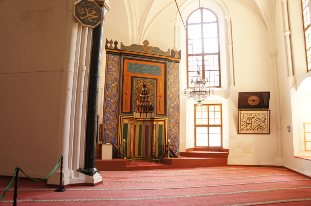 This image shows the interior of a mosque. The focal point is the mihrab, a decorated niche in the wall that indicates the direction of Mecca. The mihrab is ornately decorated with intricate patterns and calligraphy. Above it, there is a large chandelier hanging from the ceiling. The walls are adorned with Arabic calligraphy and other decorative elements. The floor is covered with red prayer rugs, and there are large windows allowing natural light to illuminate the space. The architecture features high, arched ceilings and a serene, spacious atmosphere.
