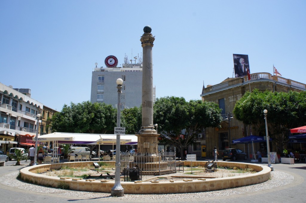 Alt text: A public square with a tall stone column in the center, surrounded by a circular fence and a small garden area. The square is bordered by buildings, including a modern multi-story building with a large clock on top and an older building with a billboard. There are trees providing shade, and several outdoor seating areas with umbrellas. People are walking and sitting around the square, enjoying the sunny day.