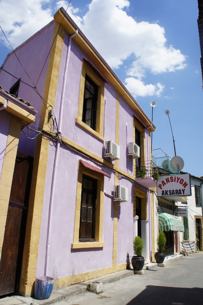 The image shows a street scene with a two-story building painted in light purple with yellow trim. The building has several windows, some with awnings, and air conditioning units attached to the exterior. There is a sign on the building that reads "Pansiyon Aksaray." In front of the building, there are potted plants. The sky is clear with a few clouds, indicating a sunny day. Other buildings and a market with a green and white striped awning are visible further down the street.