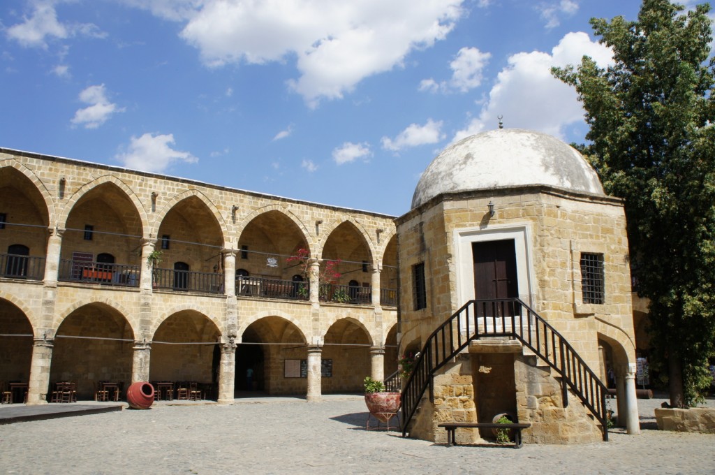 The image shows a historic stone building with arched doorways and windows, arranged in a rectangular courtyard. The building has two levels, with the upper level featuring a balcony with railings. In the center of the courtyard, there is a small, domed structure with a staircase leading up to a door. The sky is clear with a few scattered clouds, and there is a tree on the right side of the image. The courtyard appears to be paved with cobblestones, and there are a few pieces of furniture and potted plants around.