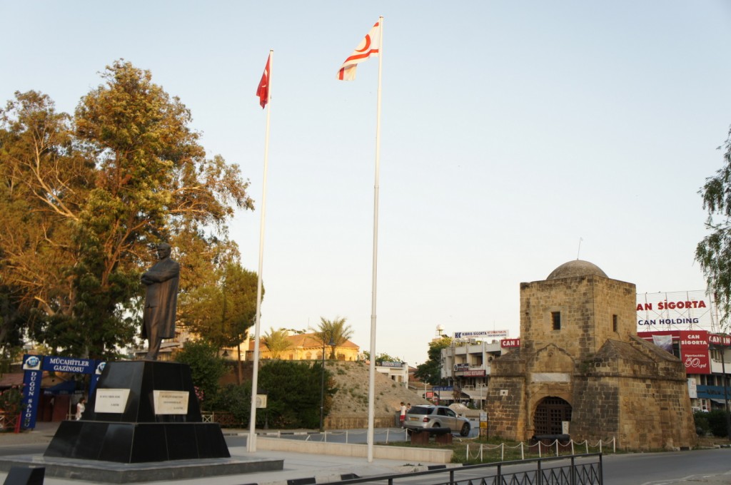 Alt text: A public square featuring a statue of a person on a pedestal, with two tall flagpoles flying flags. To the right, there is an old stone building with a domed roof. Trees and various buildings, including one with a sign that reads "CAN SİGORTA," are visible in the background. The sky is clear and the scene is well-lit, suggesting it is daytime.