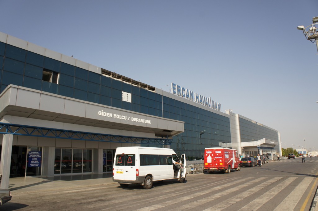 The image shows the exterior of Ercan Airport (Ercan Havalimanı) with a focus on the departure area. The building has a modern design with large glass windows. There are signs in both Turkish and English indicating the departure area. In front of the building, there are a few vehicles, including a white van and a red vehicle, parked along the curb. The sky is clear, suggesting a sunny day.