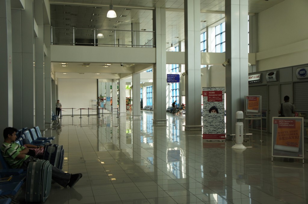 The image shows the interior of an airport terminal. The terminal has a high ceiling with large windows allowing natural light to fill the space. There are rows of blue seats on the left side, with a few people sitting and waiting. On the right side, there are closed shops with signs, including one for car rentals. In the middle of the terminal, there are informational signs and advertisements. The floor is shiny and reflective, indicating it is well-maintained. The overall atmosphere is calm and quiet.