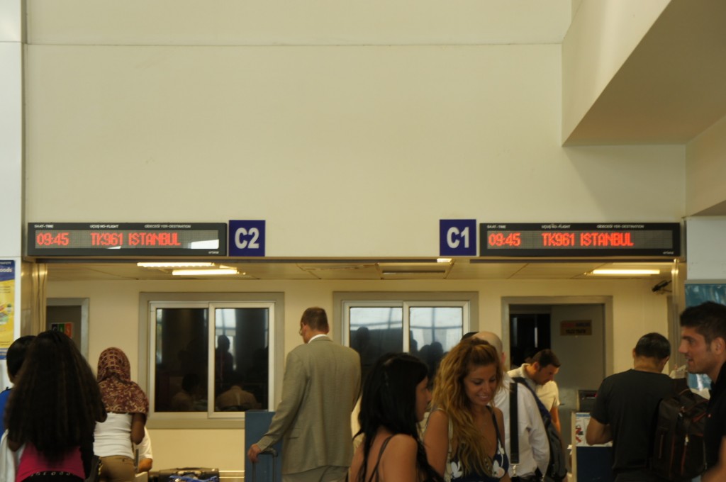 The image shows a busy airport gate area with people waiting in line. Above the gates, there are digital display boards indicating flight information. The displays show the flight number TK961 to Istanbul, with a departure time of 09:45. The gates are labeled C1 and C2. Several passengers are visible, some standing and some engaged in conversation. The atmosphere appears to be typical of an airport boarding area.