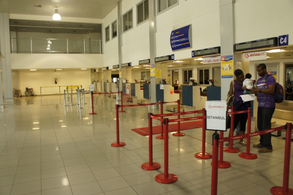 The image shows an airport check-in area with several check-in counters. There are red barriers guiding passengers to the counters, and a sign indicating a flight to Istanbul. Two people, one holding a baby, are standing near the check-in area, possibly reviewing travel documents. The area appears to be relatively empty, with only a few people visible in the background. The counters are labeled with signs for "Atlasjet" and there are overhead signs providing directions and information. The floor is tiled, and the area is well-lit with overhead lighting.