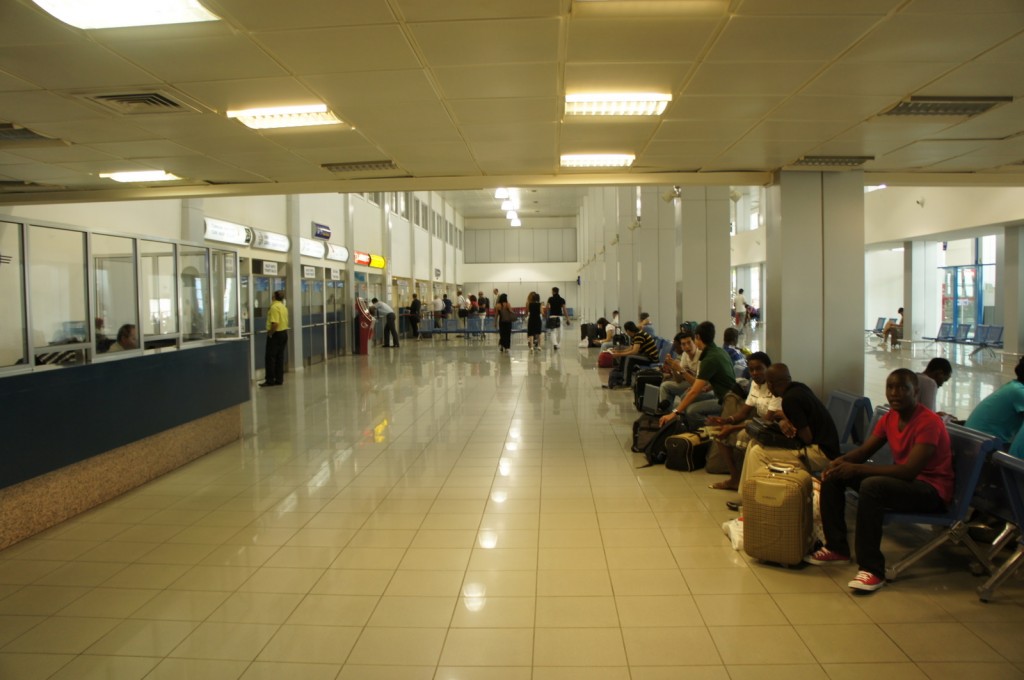 The image shows the interior of an airport terminal. On the left side, there are several service counters with glass partitions and signs above them. A few people are standing near the counters, and some staff members are visible behind the glass. On the right side, there are rows of blue chairs where passengers are seated, some with luggage beside them. The terminal has a clean, tiled floor and a high ceiling with fluorescent lighting. In the background, more passengers can be seen walking or waiting. The overall atmosphere appears to be calm and orderly.