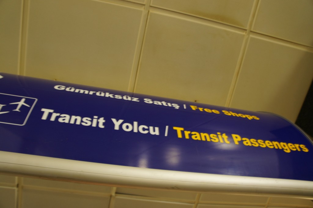 The image shows a blue airport sign with white and yellow text. The sign has the words "Gümrüksüz Satış / Free Shops" in white and yellow, and "Transit Yolcu / Transit Passengers" in white and yellow. There is also a small icon of an airplane on the left side of the sign. The sign is located on a ceiling with a tiled pattern.