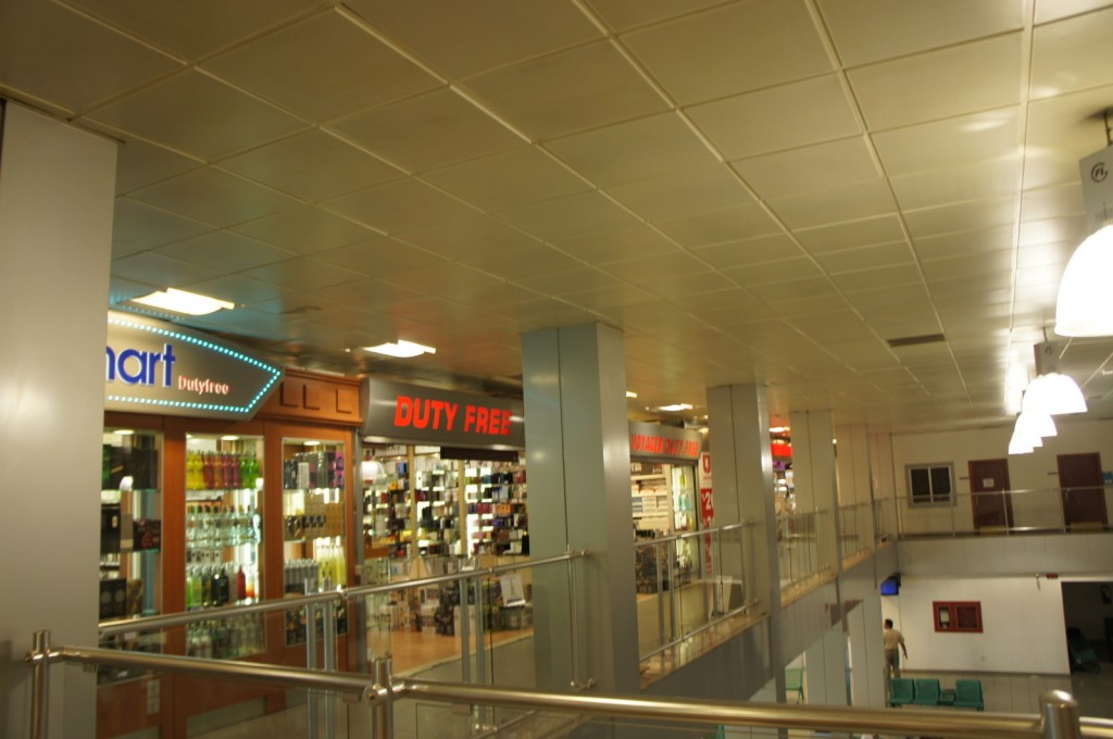 The image shows the interior of an airport or shopping mall with a focus on a duty-free shopping area. There are several stores visible, including one with a sign that reads "Duty Free" in red letters and another with a sign that reads "smart Dutyfree." The stores display various products, likely perfumes, cosmetics, and other duty-free items. The area has a modern design with a tiled ceiling, metal railings, and bright lighting. There are also some seating areas visible in the lower part of the image.