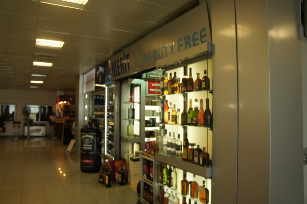 The image shows the interior of a duty-free shop at an airport. The shop has a sign that reads "MERIT elite DUTY FREE" and displays various bottles of alcohol on illuminated shelves. The store is well-lit with overhead fluorescent lights, and there are other shops and seating areas visible in the background. The floor is tiled, and the overall atmosphere suggests a clean and modern airport environment.