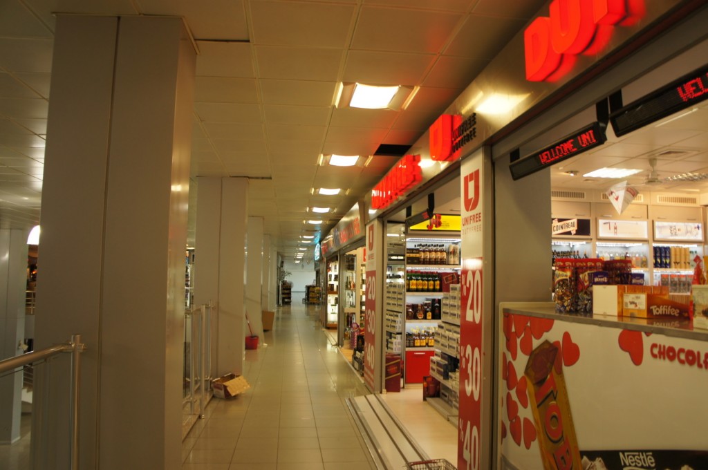 The image shows the interior of a duty-free shop in an airport or similar location. The store has bright lighting and is well-stocked with various products, including chocolates, liquor, and other items typically found in duty-free shops. There are signs with red text and promotional offers, such as "20%," "30%," and "40%." The aisle is clean and spacious, with shelves and displays on both sides. The ceiling has recessed lighting, and there are structural columns along the walkway.