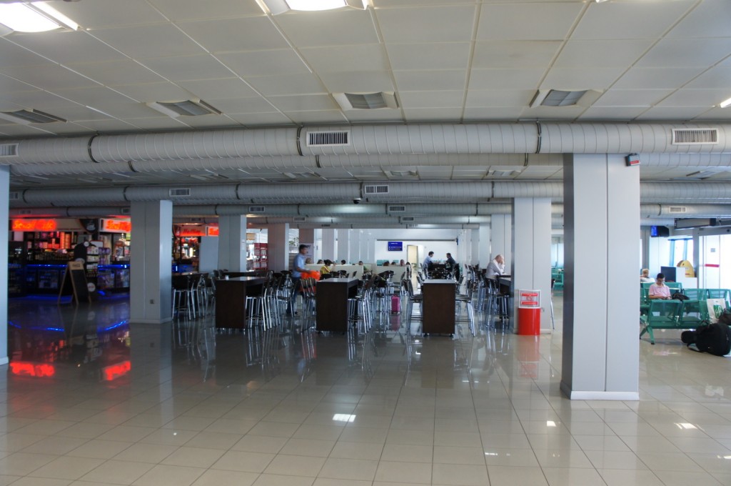 The image shows a spacious indoor area, likely an airport terminal. The space has a high ceiling with visible air ducts and is well-lit with fluorescent lights. There are several seating areas with tables and chairs, some of which are occupied by people. On the left side, there is a bar or café with illuminated signs and a person standing behind the counter. The floor is tiled and shiny, reflecting the lights and furniture. In the background, there are more people seated, and some are walking around. There is also a red trash bin near one of the columns.
