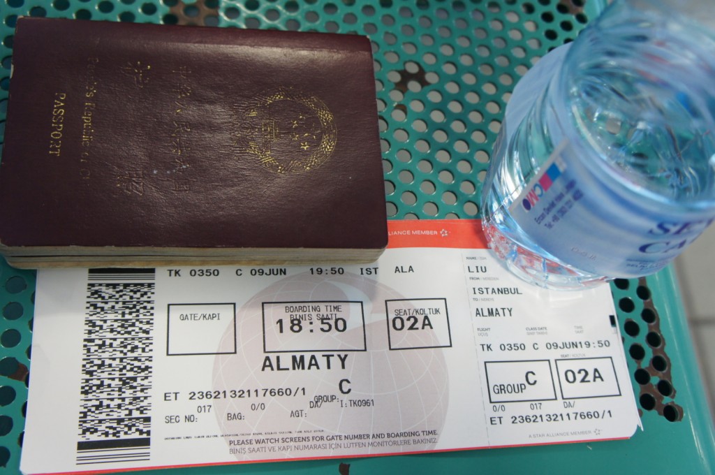 The image shows a close-up of a traveler's items on a perforated metal surface. There is a maroon-colored passport, a boarding pass, and a bottle of water. The boarding pass indicates a flight from Istanbul to Almaty with the flight number TK 0350, departing on June 9th at 19:50. The seat number is 02A, and the passenger's name is partially visible as "LIU."
